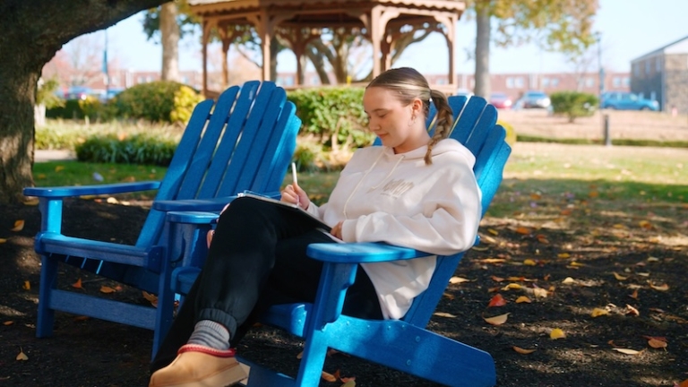 Student sitting in blue Adirondack chair on campus with a gazebo in the background